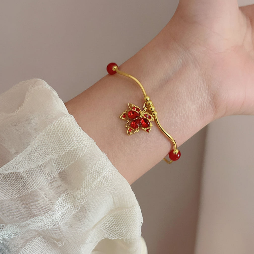 A gold-toned bracelet with red gemstone accents and a floral motif, shown worn on a person's wrist with a white sheer sleeve visible.