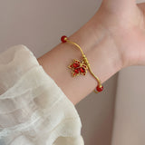 A gold-toned bracelet with red gemstone accents and a floral motif, shown worn on a person's wrist with a white sheer sleeve visible.